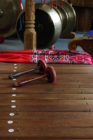 Two wooden and red beaters on a wooden xylophone, with a decorative piece of red cloth and some metal gongs in the background.