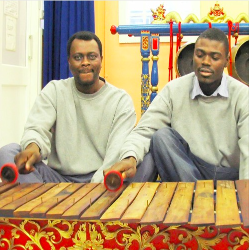 Two young black men in grey sweatshirts playing a wooden xylophone together, as part of a gamelan orchestra.