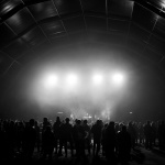 Black and white photograph of a large standing audience in a huge, spacious venue that looks like a warehouse.